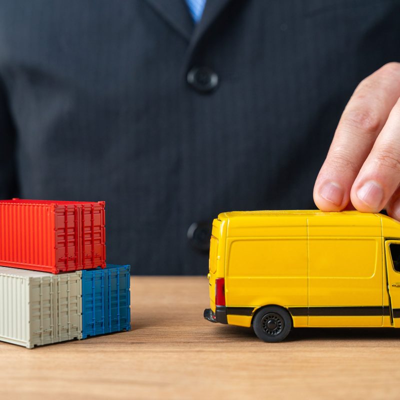 A businessman pushes a delivery van past a stack of shipping containers. The concept of delivery and transportation of goods and parcels to the end customer. Logistics and speed.